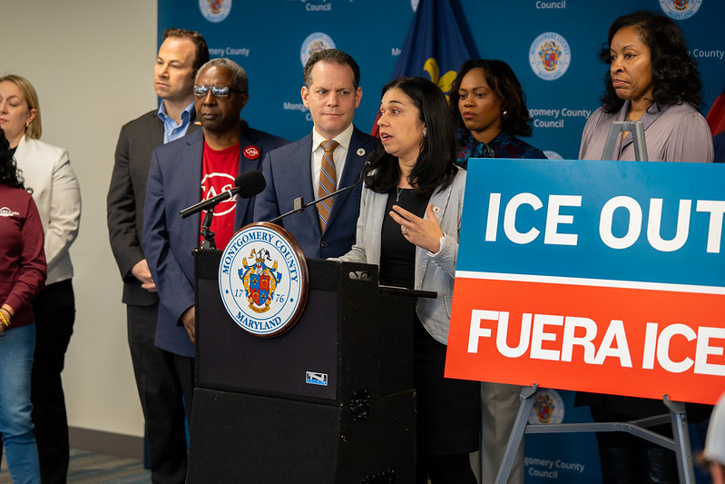 Council President Fani-González speaks at a podium next to a poster board reading “ICE OUT: FUERA ICE”. Councilmembers and community members stand beside her.