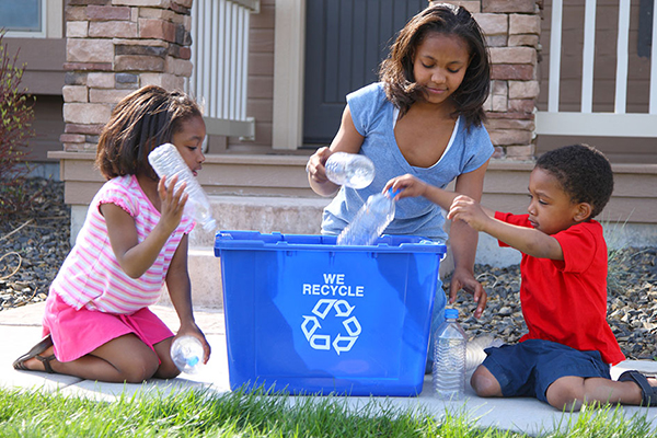 A mother and two daughters placing empty plastic water bottles into blue recycling bin in the front yard of single-family home.