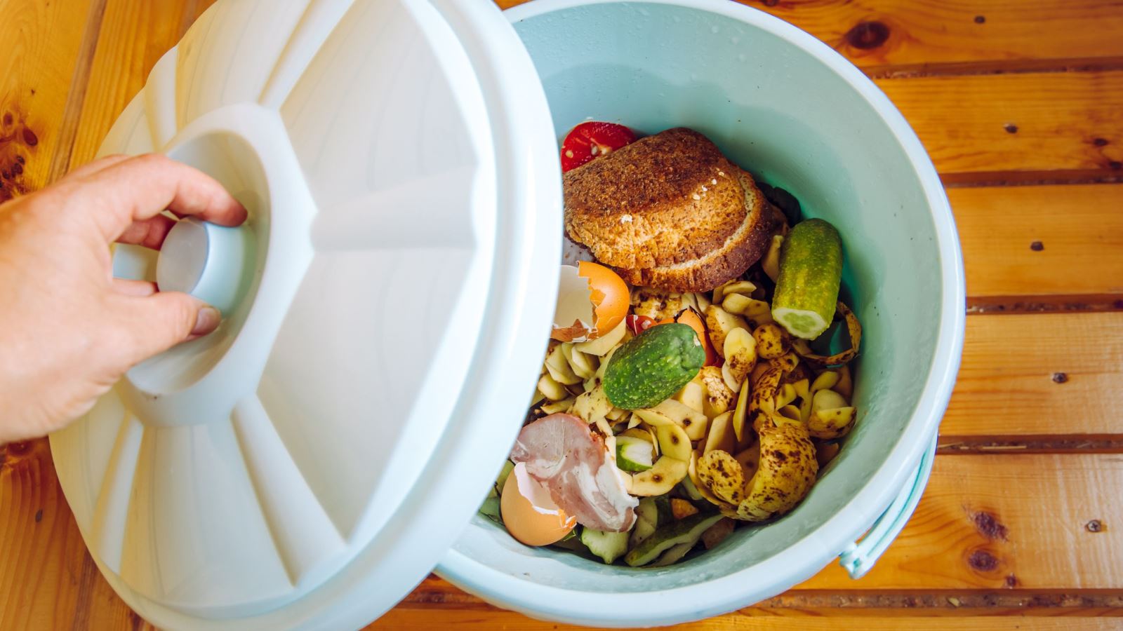 Person lifting the lid of a white ceramic container filled with food scraps. The container is on a wooden butcher countertop.