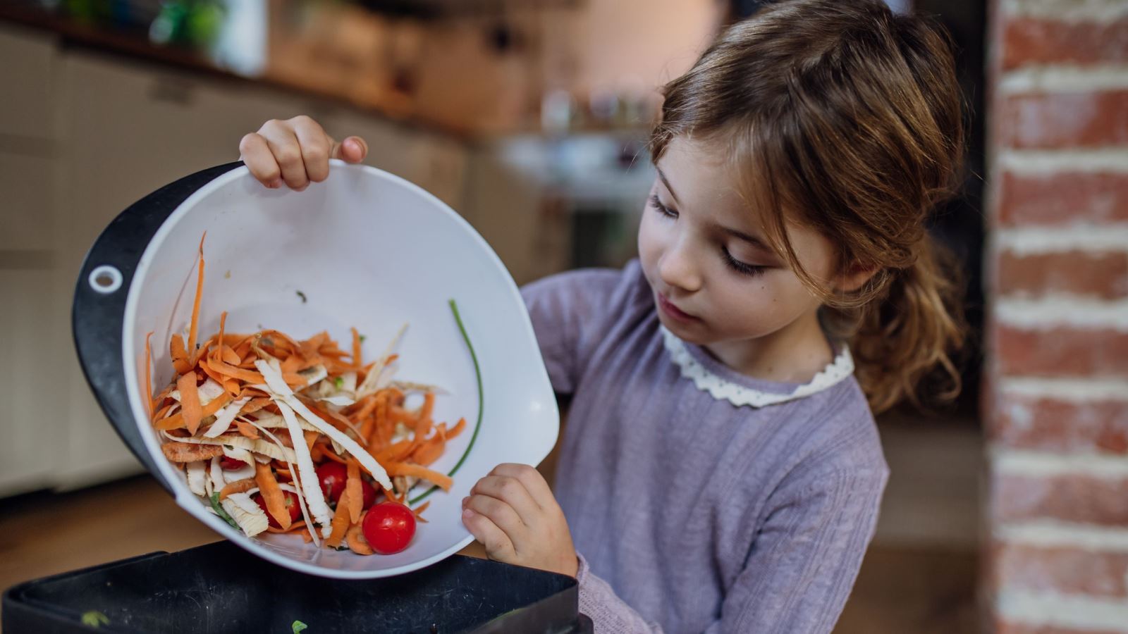 A young child tipping a bowl of vegetable peelings and whole tomatoes into the kitchen composting bin.
