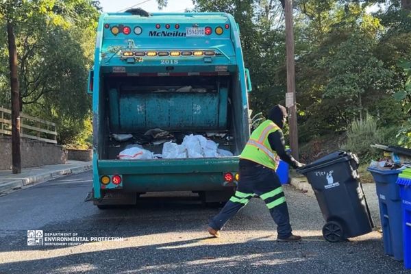 Trash collector moving black trash container off of curb. Behind him is the open end of a blue trash truck.