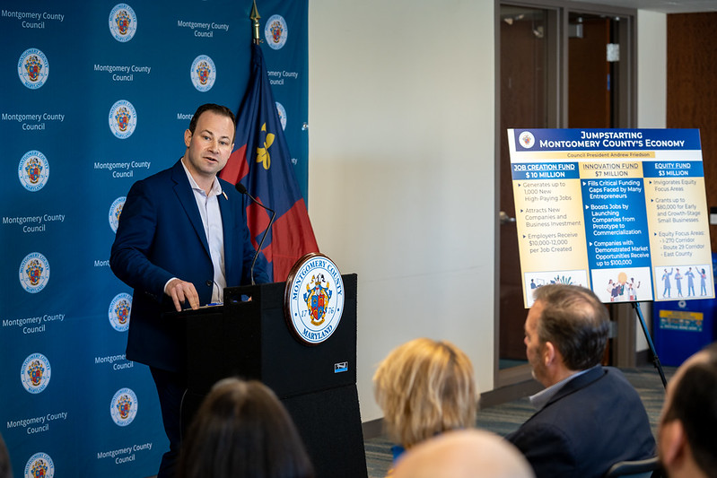 Presidente del Concejo Andrew Friedson habla en un podio al lado de un cartel que dice “Reactivemos la Economía del Condado de Montgomery” en inglés “Jumpstarting Montgomery County’s Economy”.