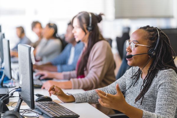 Call center operators at their computers wearing headsets out of focus. The operator in focus is wearing glasses and speaking with a caller.