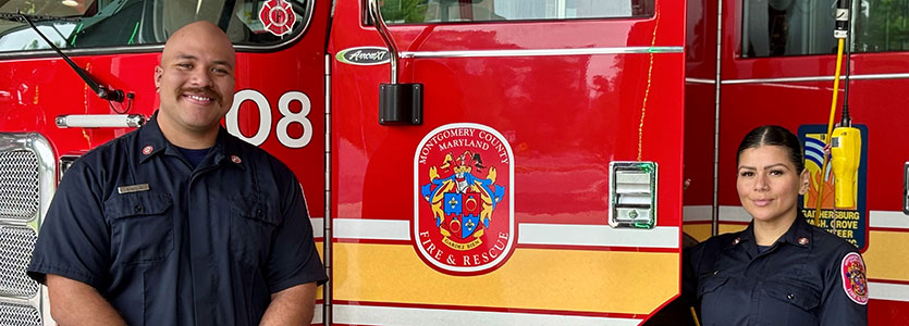 Two firefighters in uniform standing in front of the driver's side door of a fire truck